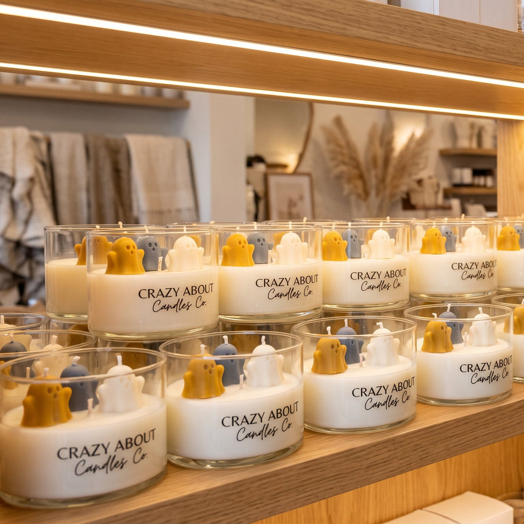 Multiple glass jar candles with white wax and gold wicks arranged on warm wooden shelves in a retail or home goods store setting.
