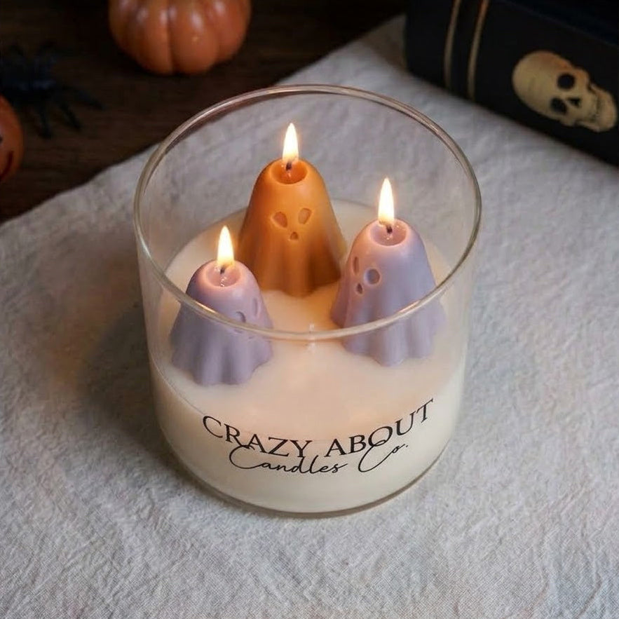 A glass jar candle with three ghost-shaped wax candles inside, lit and set on a textured white cloth. The jar is branded &