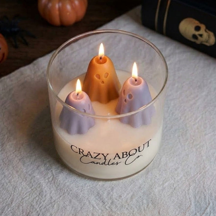 A glass jar candle with three ghost-shaped wax candles inside, lit and set on a textured white cloth. The jar is branded &