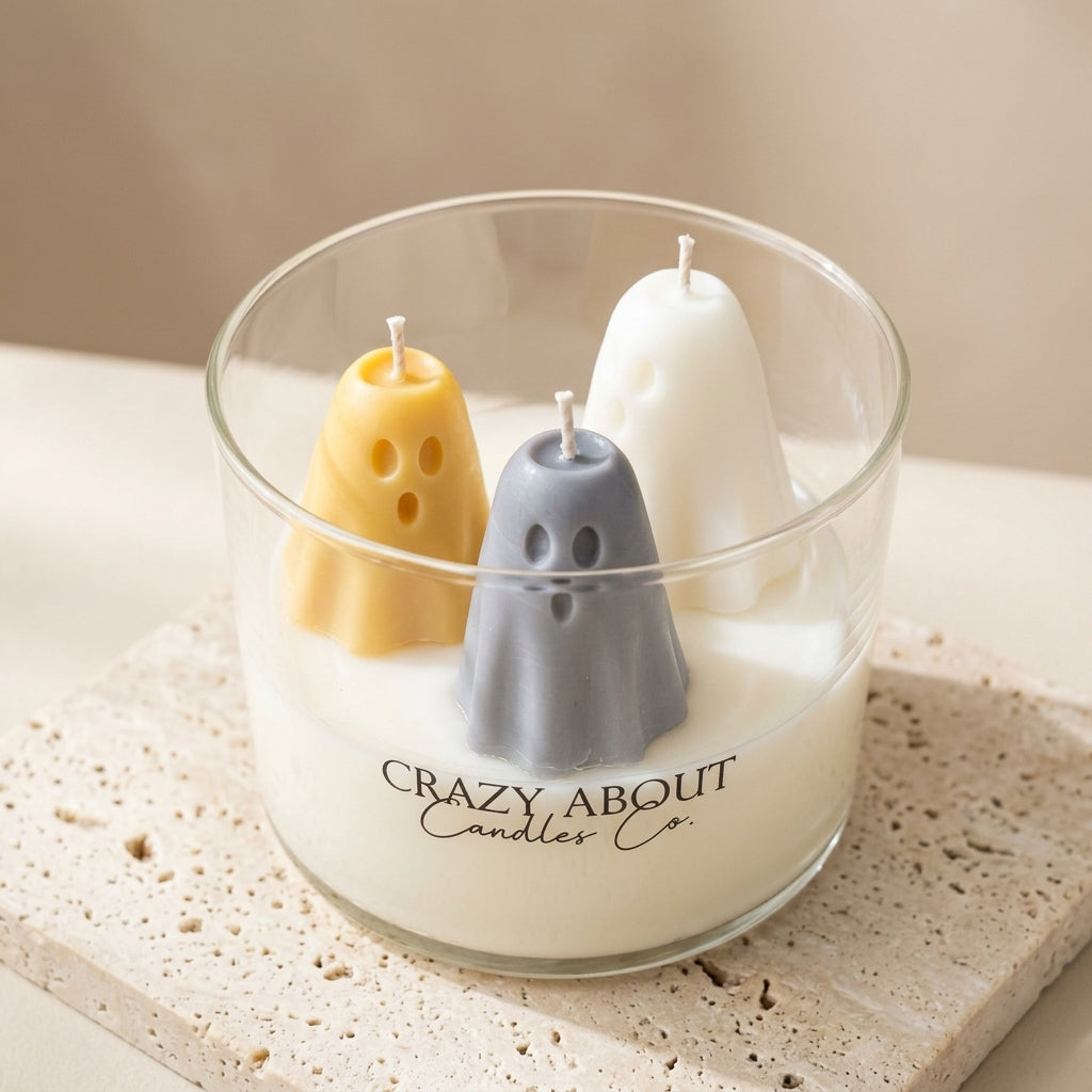 A glass jar candle with a white wax base and three ghost-shaped candles inside: one yellow, one gray, and one white. The glass jar has the text 'CRAZY, ABOUT Candles Co.