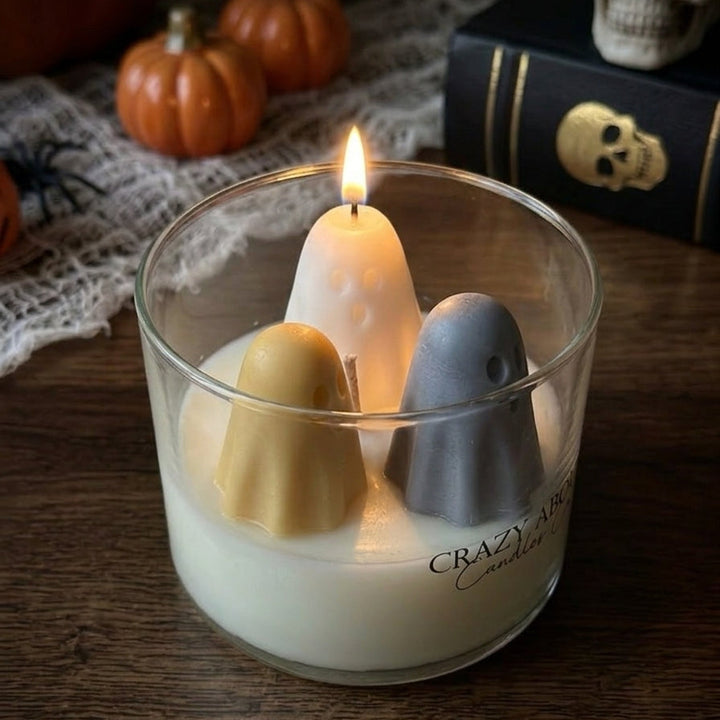 A Halloween-themed glass jar candle with three ghost-shaped wicks in beige, white, and gray, lit with a warm flame. The candle is set on a dark wooden table with seasonal props including miniature pumpkins, a skull, and a black book in the background.