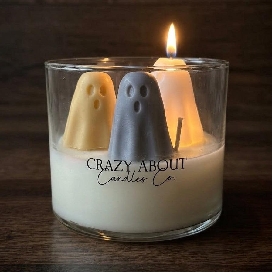 A glass jar candle with white wax and two ghost-shaped candles floating inside, one in pale yellow and one in light gray, with one ghost candle lit and burning. The glass is printed with the brand name 'Crazy About Candles Co.
