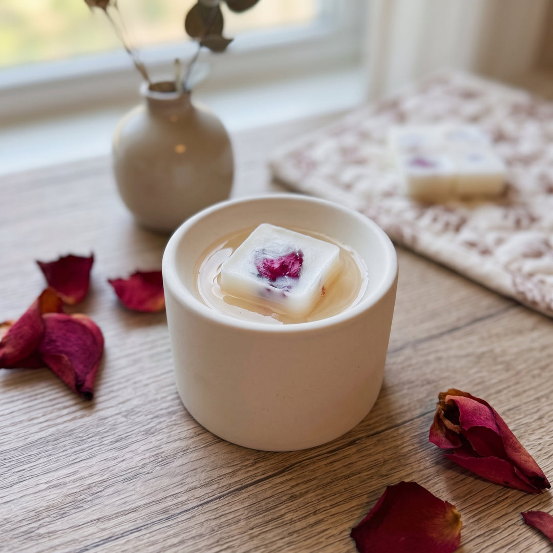 A small white ceramic cup on a wooden table holding a square white soap block with a pressed pink flower inside. The scene is styled with dried rose petals, a small vase, and another soap bar on a cloth in the background.
