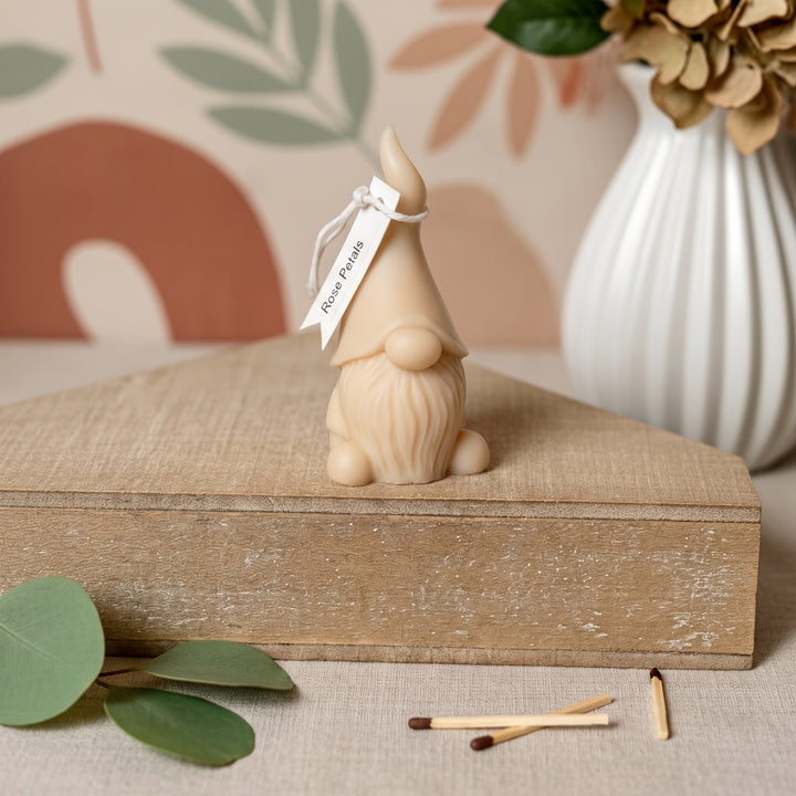 A small beige gnome figurine with a pointed hat and long beard sits on a rustic wooden box. A white tag tied to the hat reads &