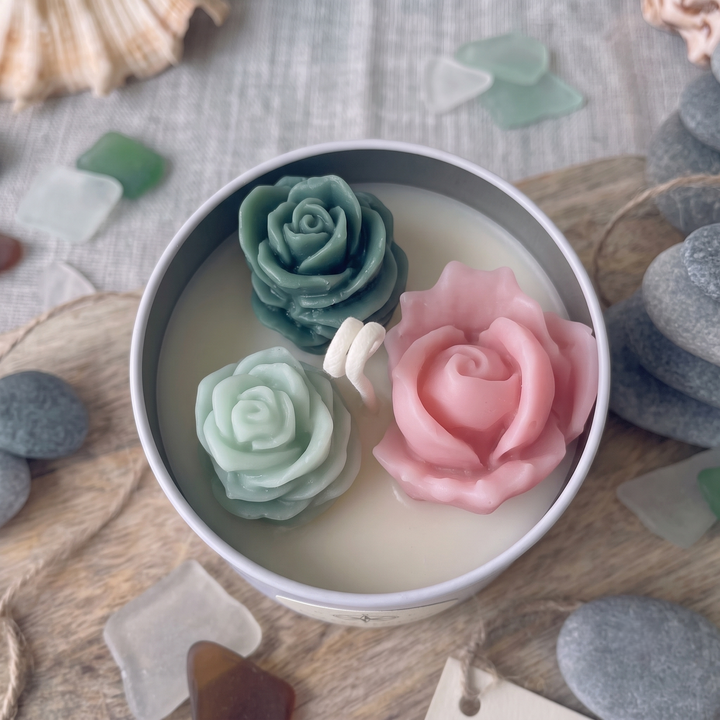 A round tin candle with three sculpted rose-shaped wax inserts in teal, pale mint, and soft pink, styled on a wooden surface with sea glass and stones.