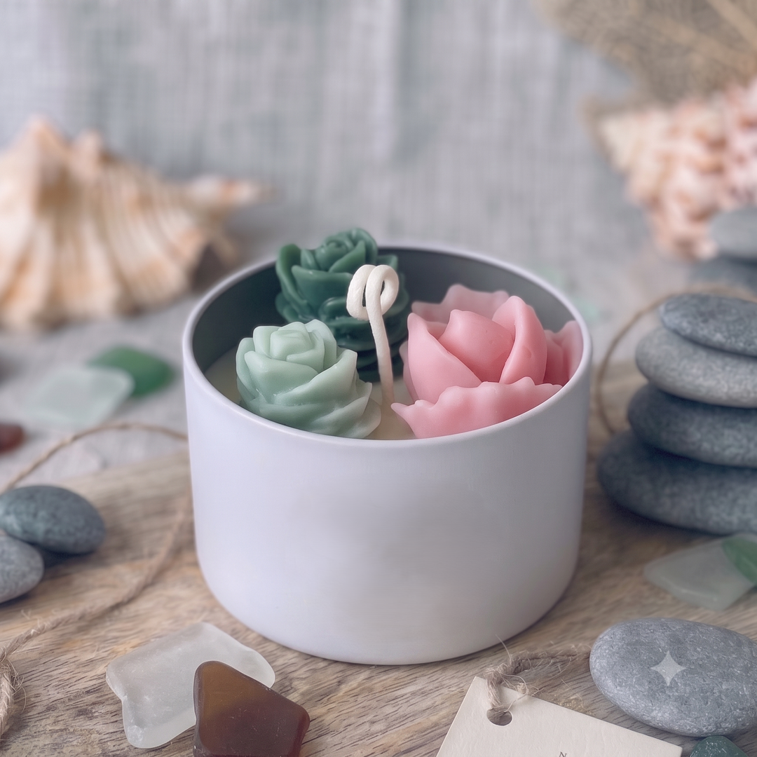 A white ceramic candle holder filled with sculpted rose-shaped wax candles in pastel green, pink, and cream, styled on a wooden surface with seashells, smooth stones, and decorative tags.