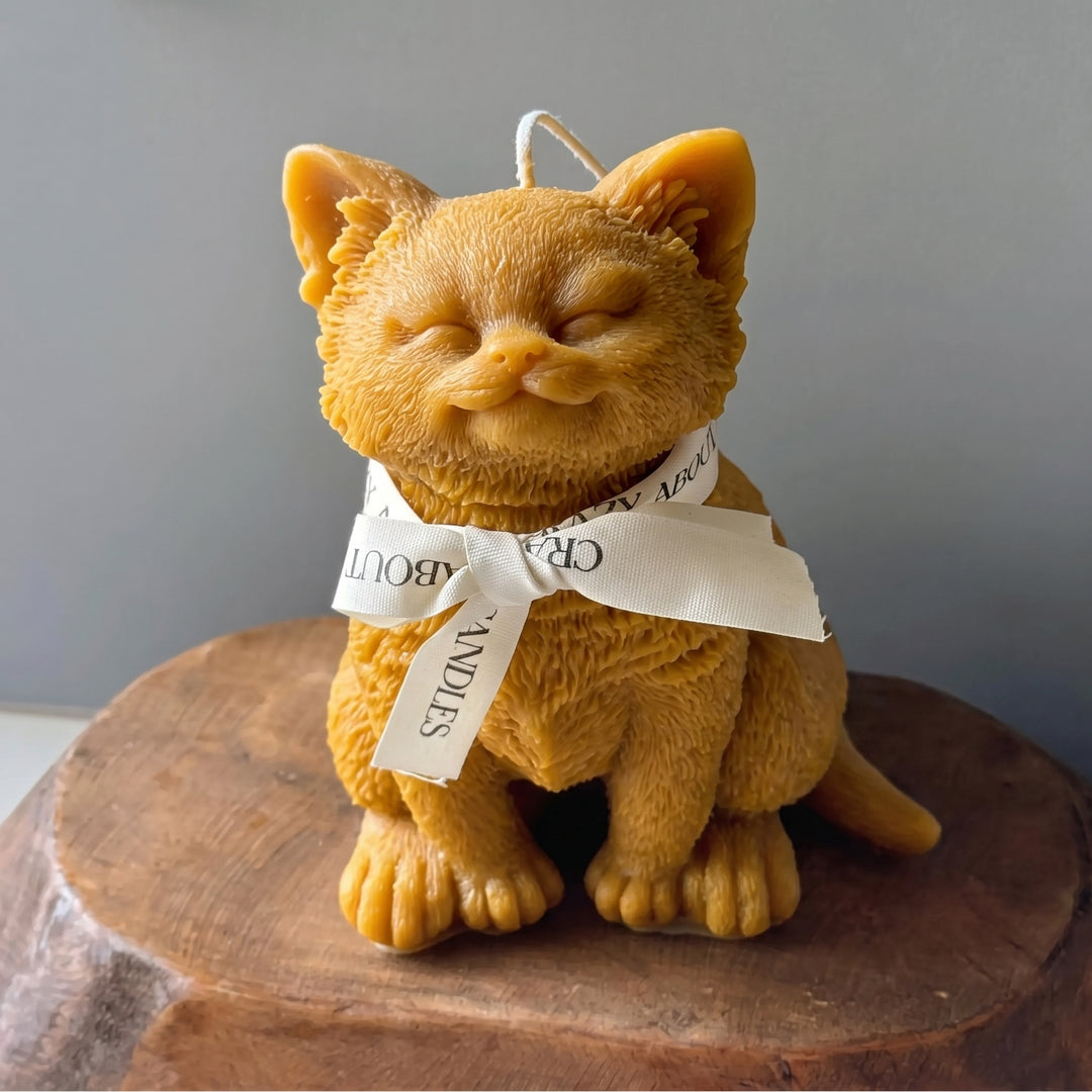A golden-yellow cat-shaped candle with closed eyes, sitting on a rustic wooden block against a neutral gray background. The candle is tied with a white ribbon that reads &