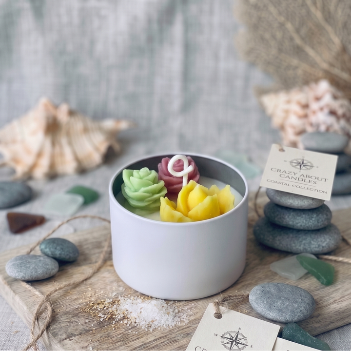 Candle set with decorative candles in a white container on a wooden surface with seashells and stones.