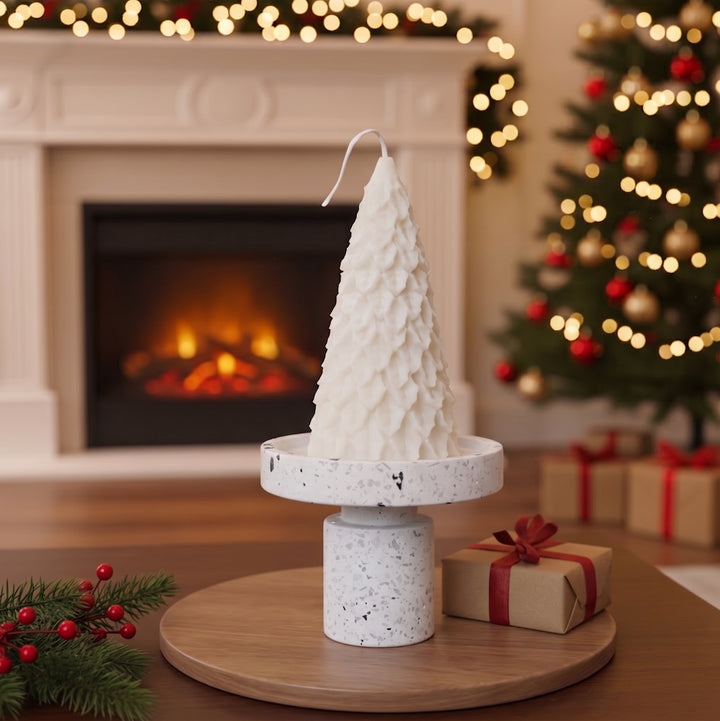 A white textured Christmas tree-shaped candle displayed on a round wooden stand, set in a cozy living room with a fireplace, a lit Christmas tree, and wrapped gifts in the background.