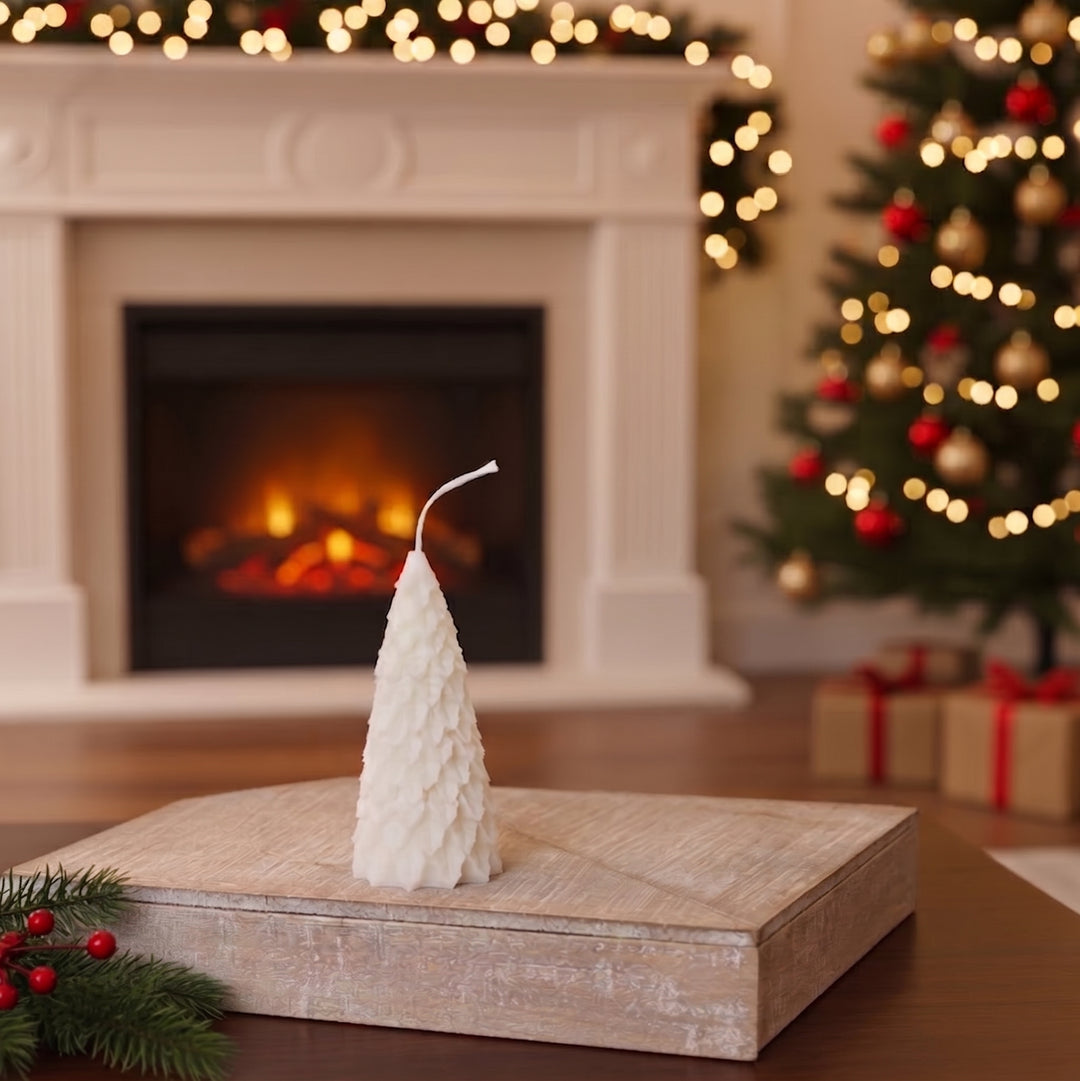 A white small pine Christmas tree-shaped candle sits on a rustic wooden box in a cozy holiday living room setting with a fireplace, decorated Christmas tree, and gifts in the background.