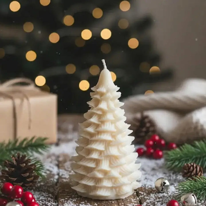 A white, tiered Christmas tree candle stands on a rustic wooden surface surrounded by holiday decorations, including pine branches, red berries, pinecones, silver bells, and a soft beige knit blanket, with a blurred Christmas tree and warm string lights in the background.