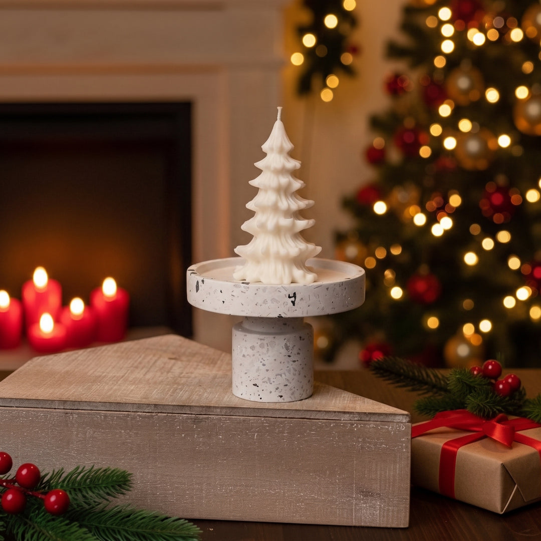 A white Christmas tree-shaped candle displayed on a speckled round pedestal stand, set on a wooden box in a cozy holiday scene with a blurred Christmas tree, fireplace candles, and wrapped gifts in the background.