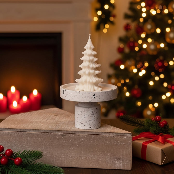 A white Christmas tree-shaped candle displayed on a speckled round pedestal stand, set on a wooden box in a cozy holiday scene with a blurred Christmas tree, fireplace candles, and wrapped gifts in the background.