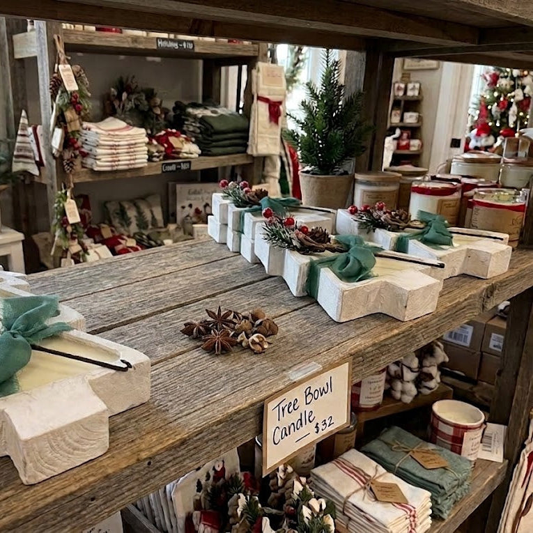 A rustic wooden table in a home decor store displays white carved Christmas tree ornaments with green bows, pinecones, and red berries. A small sign reads &