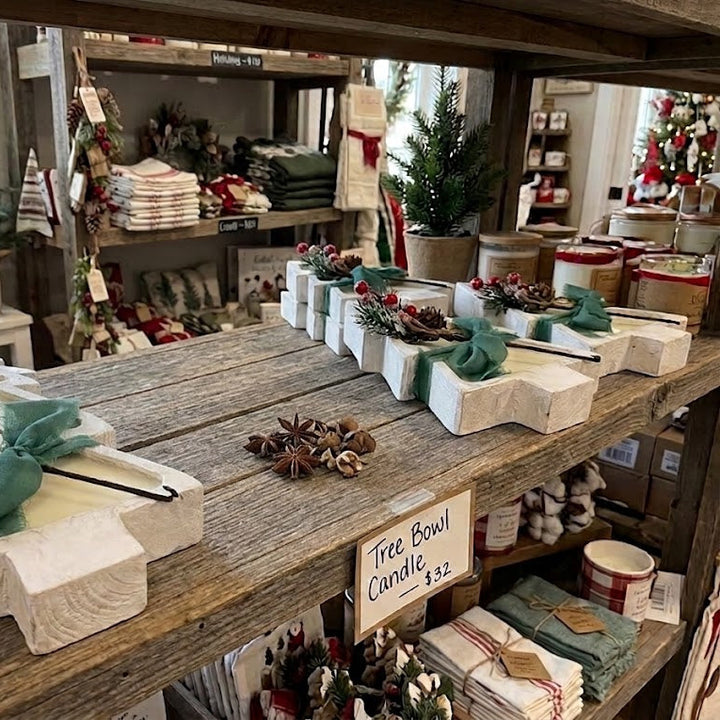 A rustic wooden table in a home decor store displays white carved Christmas tree ornaments with green bows, pinecones, and red berries. A small sign reads &