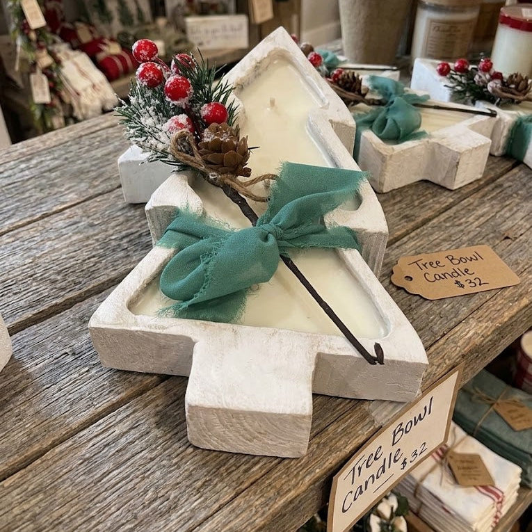 A Christmas tree-shaped wooden candle holder filled with white wax, decorated with a teal bow, greenery, pinecones, and red berries, displayed on a rustic wooden table in a gift shop.
