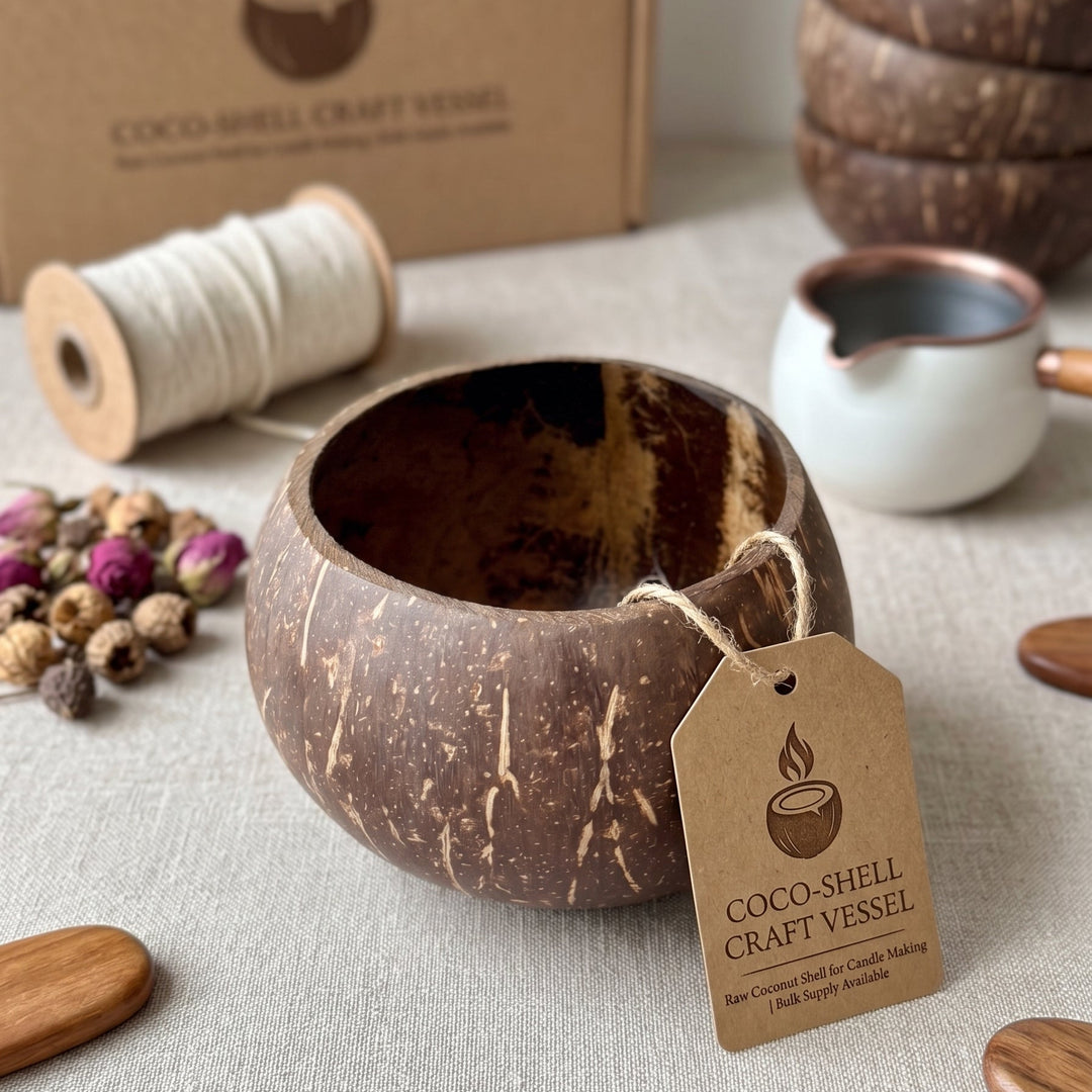 A coconut shell craft vessel with a rustic brown and black textured exterior, shown with its kraft paper tag and branded cardboard box. The vessel is styled on a neutral fabric surface with decorative props including a spool of white thread, dried flowers, wooden spoons, and a small white pitcher.