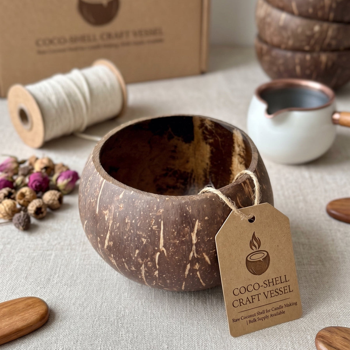 A coconut shell craft vessel with a rustic brown and black textured exterior, shown with its kraft paper tag and branded cardboard box. The vessel is styled on a neutral fabric surface with decorative props including a spool of white thread, dried flowers, wooden spoons, and a small white pitcher.