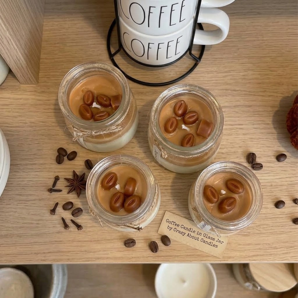 Three small glass jar candles with caramel-colored wax and coffee bean decorations are arranged on a light wood surface, with scattered coffee beans, star anise, and a small paper tag. A white ceramic mug with &