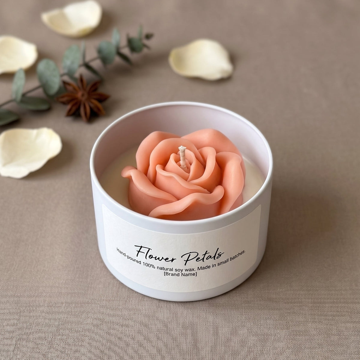 A white candle tin with a sculpted peach-pink rose wax flower on top, labeled 'Flower Petals,' set on a beige fabric surface with scattered petals and greenery in the background.