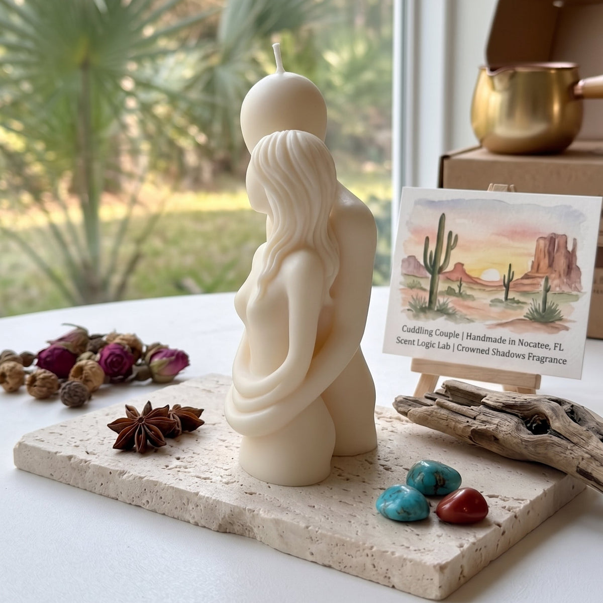 A cream-colored sculptural candle shaped like a pregnant woman cradling a fetus, displayed on a stone slab with dried flowers, turquoise stones, and a small desert-themed card in a bright indoor setting.