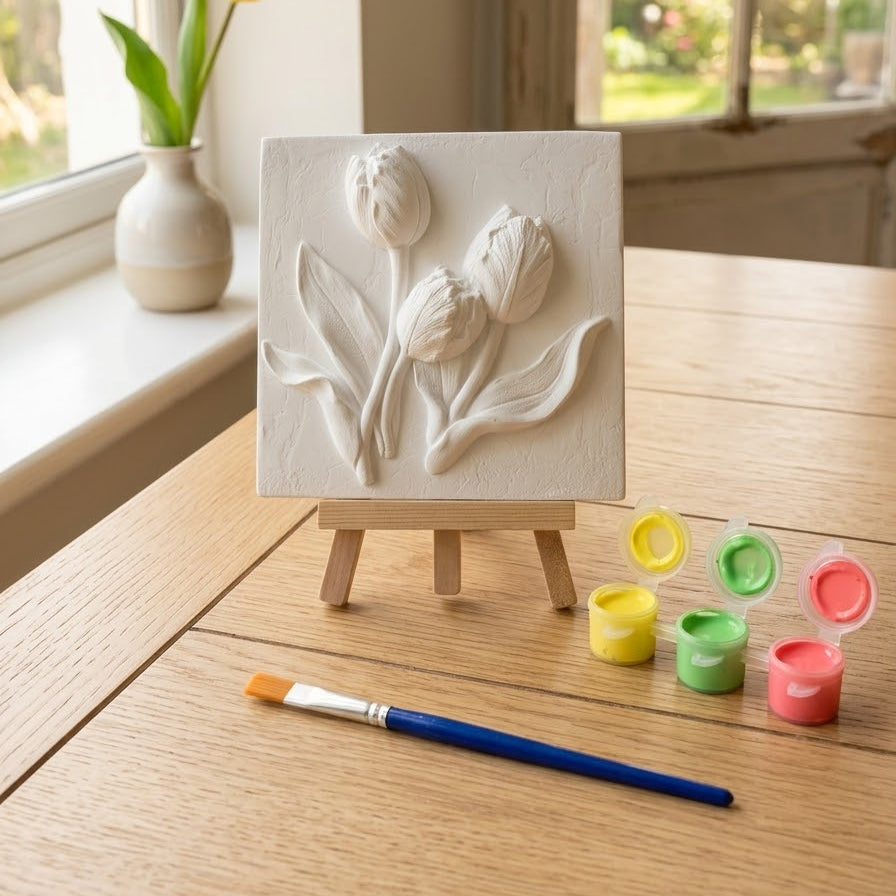 A small square canvas painting of three white tulips on a wooden easel, set on a light wood table with a paintbrush and small paint pots nearby, with a window and tulip in the background.