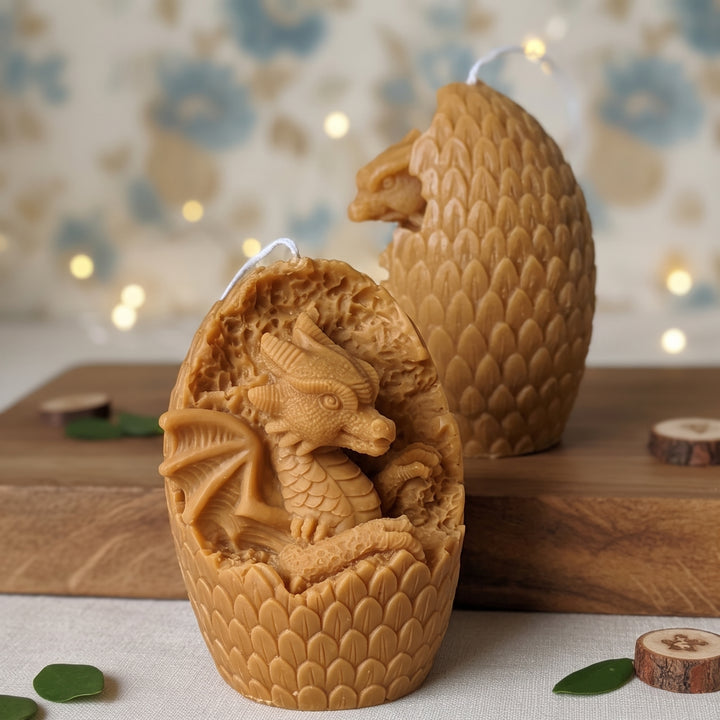 Two dragon-shaped candles carved inside textured egg-like shells, displayed on a wooden board with small green leaves and wood slices against a softly blurred floral background with warm lights.