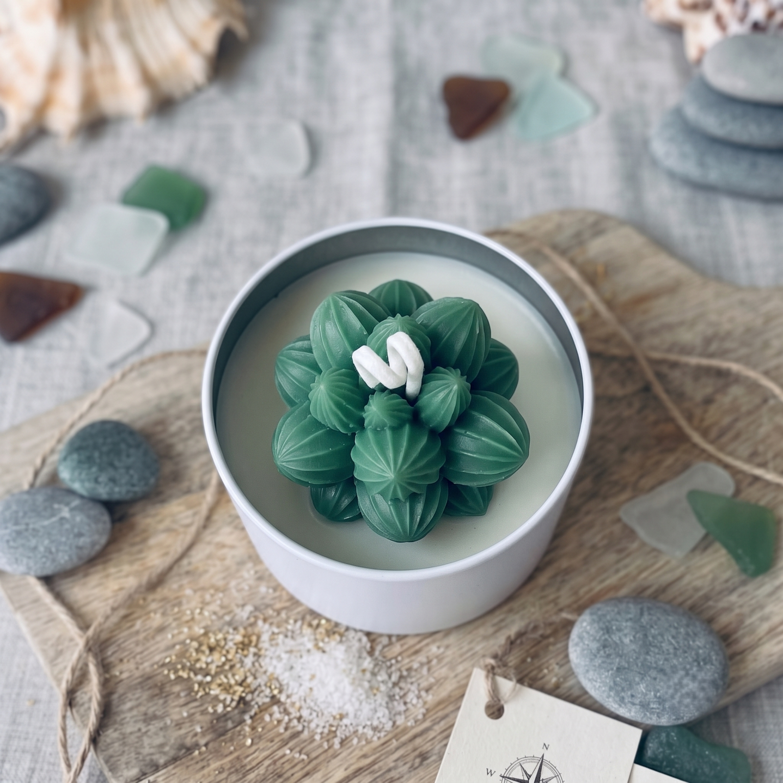 A white ceramic candle tin filled with a sculpted green succulent-shaped wax candle, styled on a wooden board with sea glass, shells, and twine.