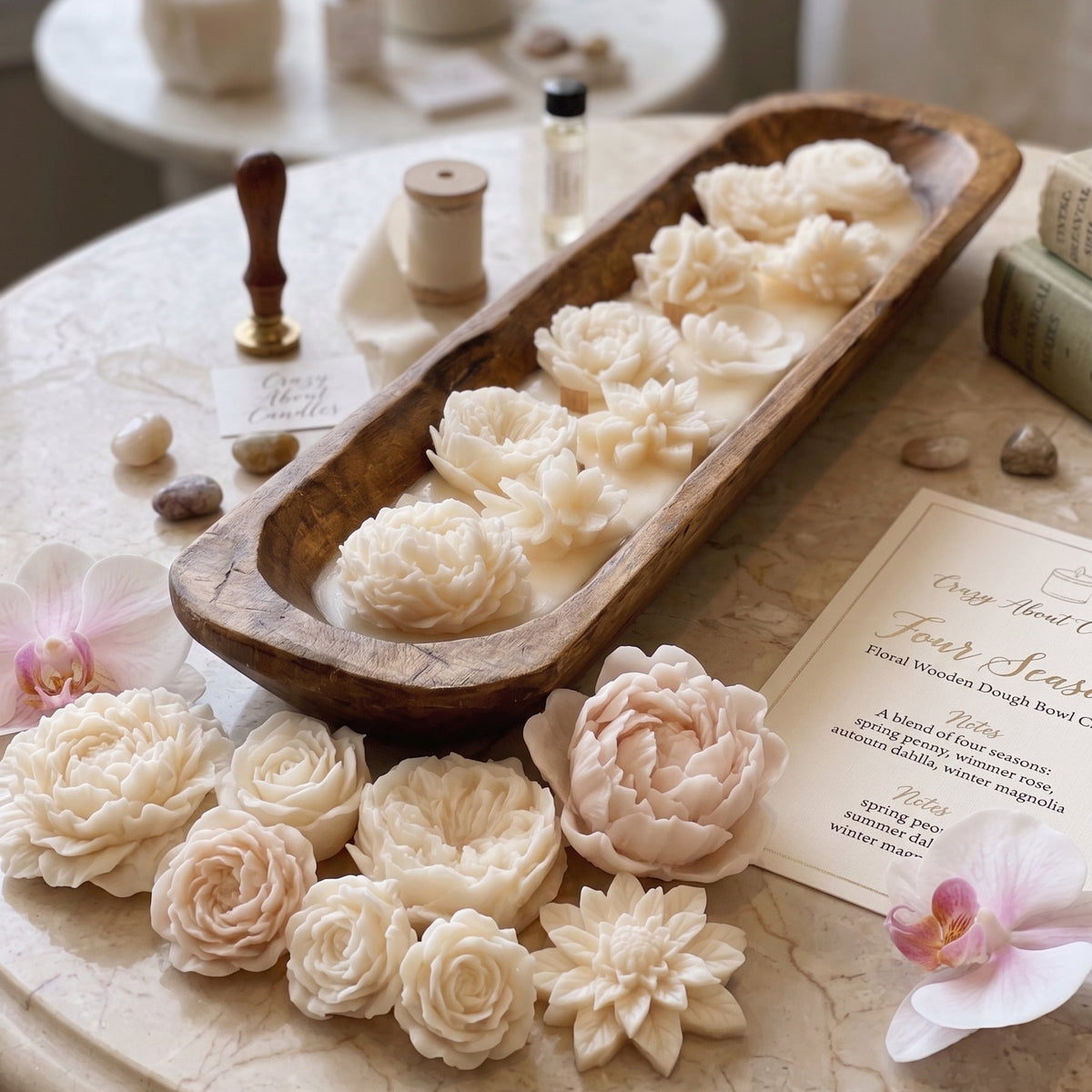 A styled tabletop display featuring multiple sculpted wax flowers in cream, ivory, and pale blush tones, arranged in a long wooden tray with additional wax flowers placed around it. The scene includes candles, small bottles, stones, a stamp, and a printed card on a marble surface near a window.