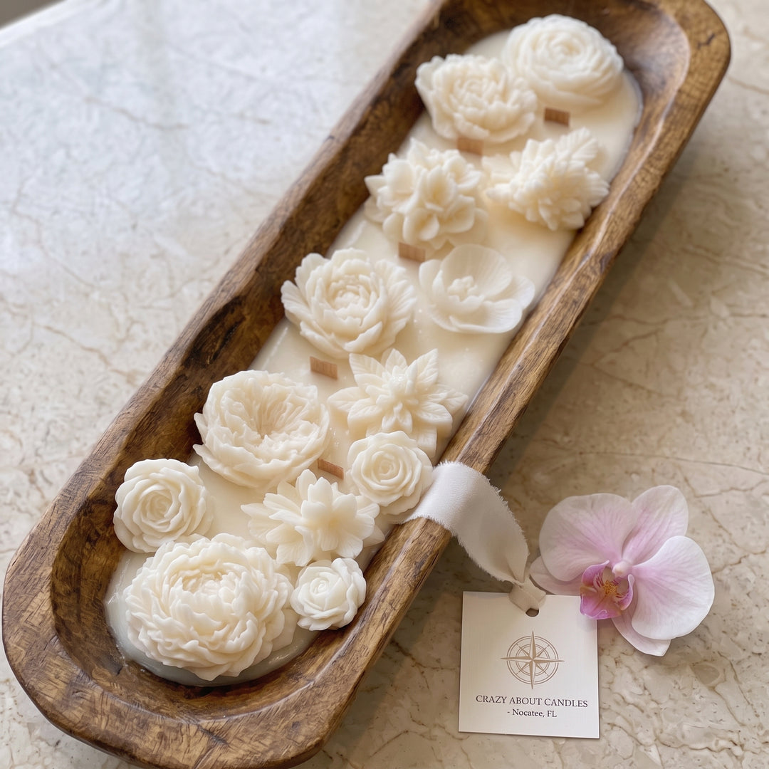 A decorative candle in a long wooden tray, featuring sculpted cream-colored floral shapes on a wax base. A small paper tag reading &