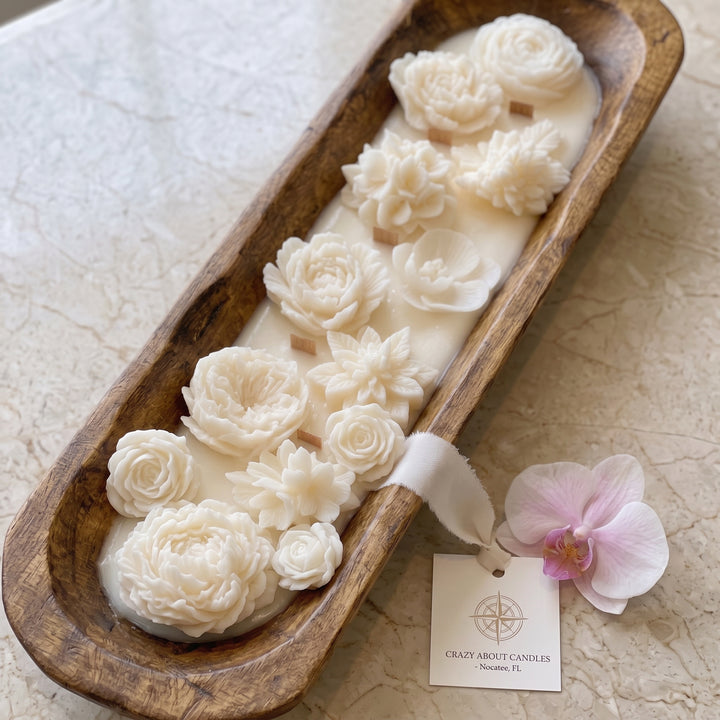 A decorative candle in a long wooden tray, featuring sculpted cream-colored floral shapes on a wax base. A small paper tag reading &