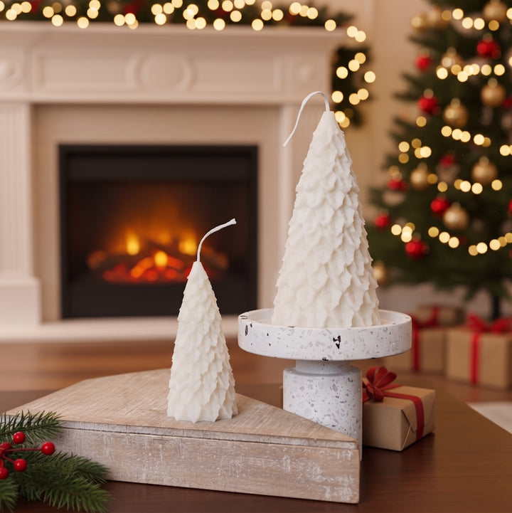 Two white textured Christmas tree-shaped candles displayed on a rustic wooden board and a white pedestal stand in a cozy holiday living room setting with a fireplace and a decorated Christmas tree in the background.
