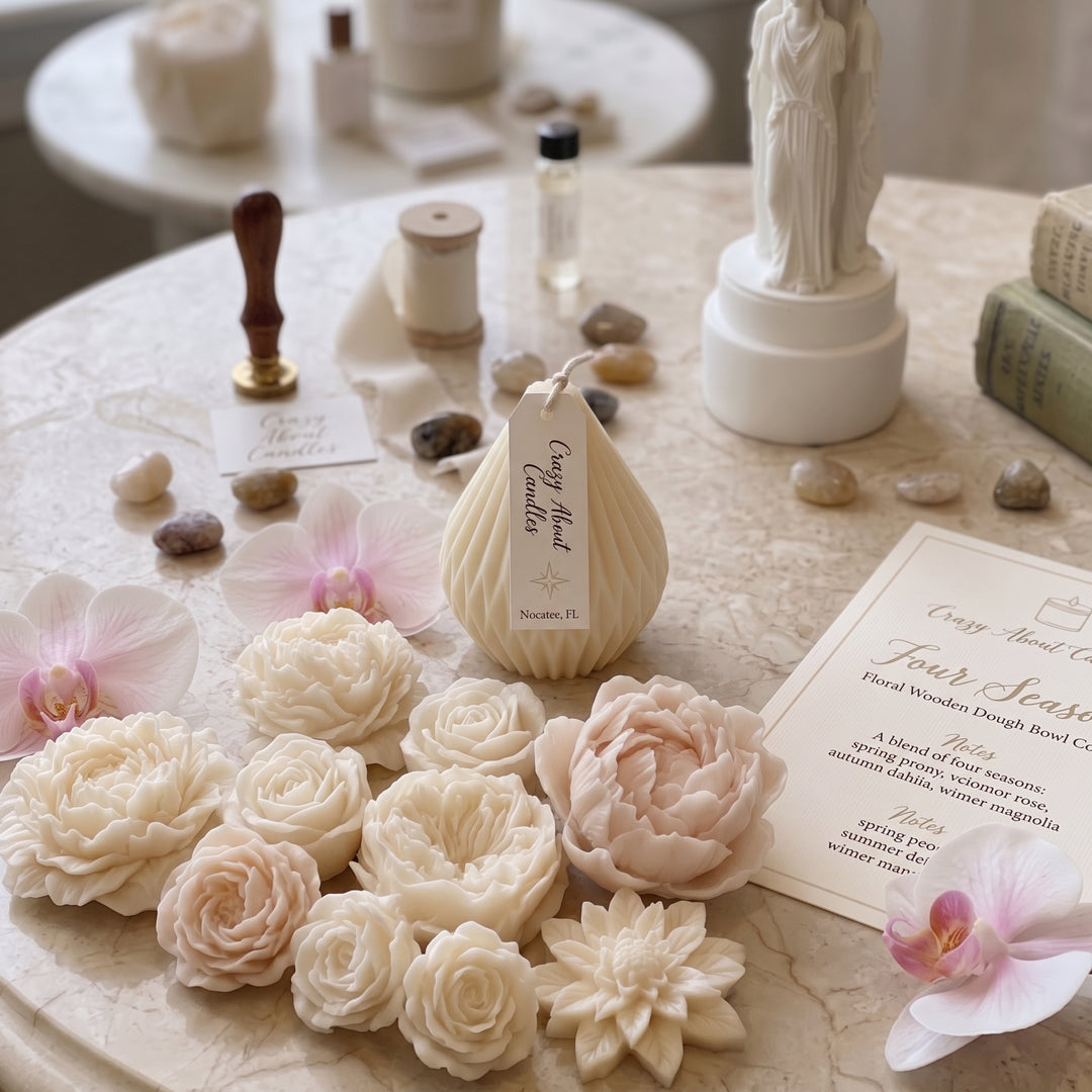 A styled tabletop display featuring a sculpted cream-colored candle shaped like a pear, surrounded by floral wax melts in cream and blush pink, crystals, a stamp, a spool of ribbon, and a printed card on a marble surface.