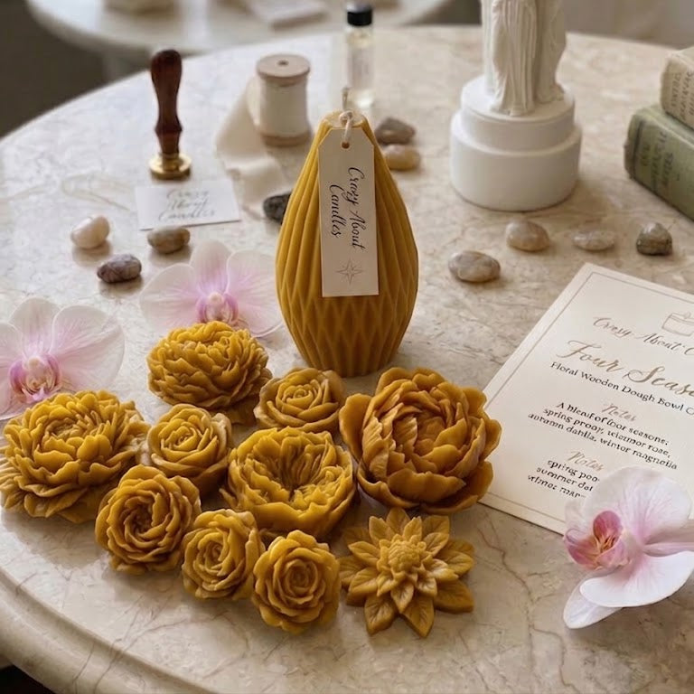 A styled tabletop display featuring a mustard yellow honeycomb candle as the main product, surrounded by wax flowers, candles, a white statue candle holder, stones, and printed cards on a marble surface.