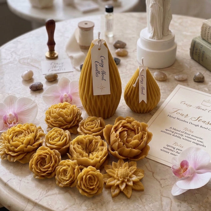 A styled tabletop display featuring handmade beeswax candles shaped like flowers and teardrop pears in warm golden yellow and amber tones, arranged with wax melts, a white statue candle holder, crystals, and printed cards on a marble surface near a window.