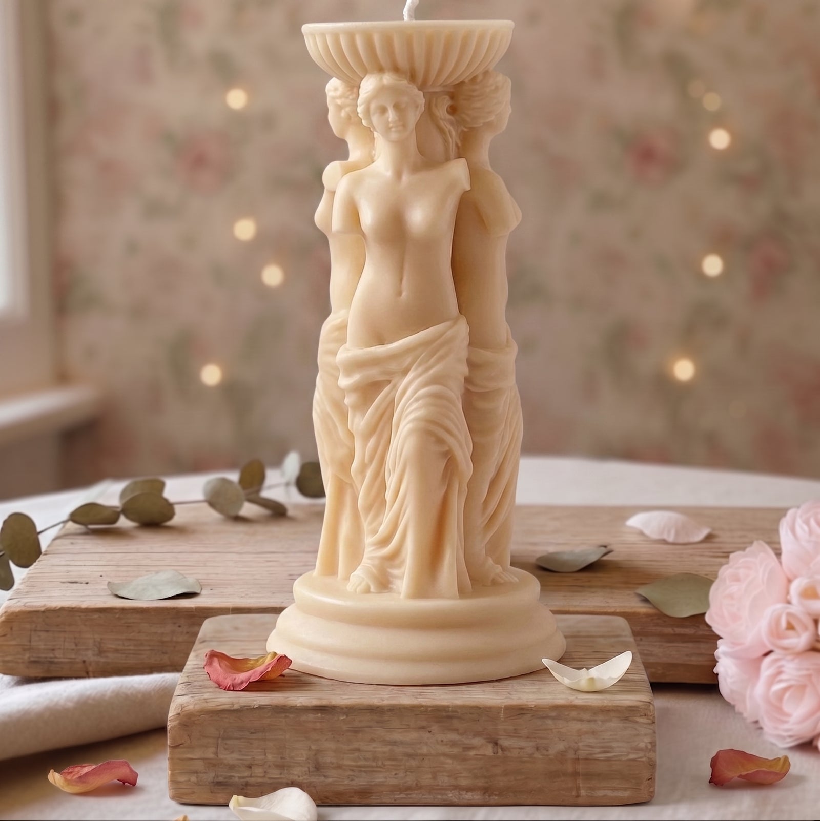 A cream-colored candle sculpted as a classical female figure standing on a pedestal, displayed on a rustic wooden board with rose petals and pink roses in a softly blurred indoor setting.