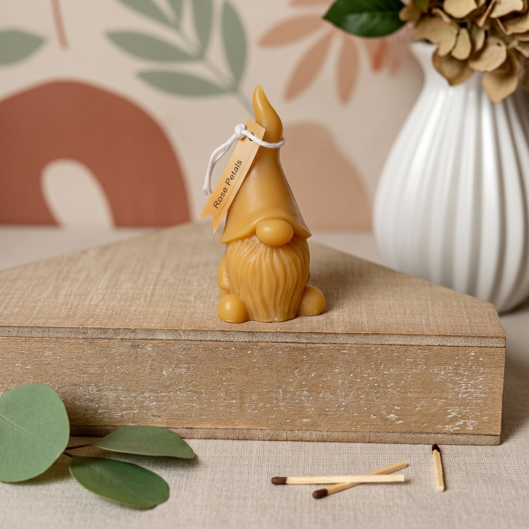 A small decorative gnome figurine in a warm tan color sits on a rustic wooden box. The figurine has a pointed hat, long beard, and a small hanging tag that reads &