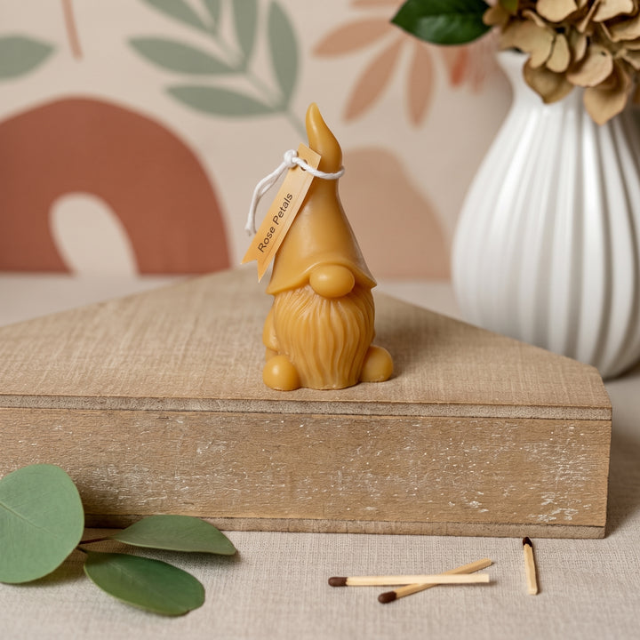 A small decorative gnome figurine in a warm tan color sits on a rustic wooden box. The figurine has a pointed hat, long beard, and a small hanging tag that reads &