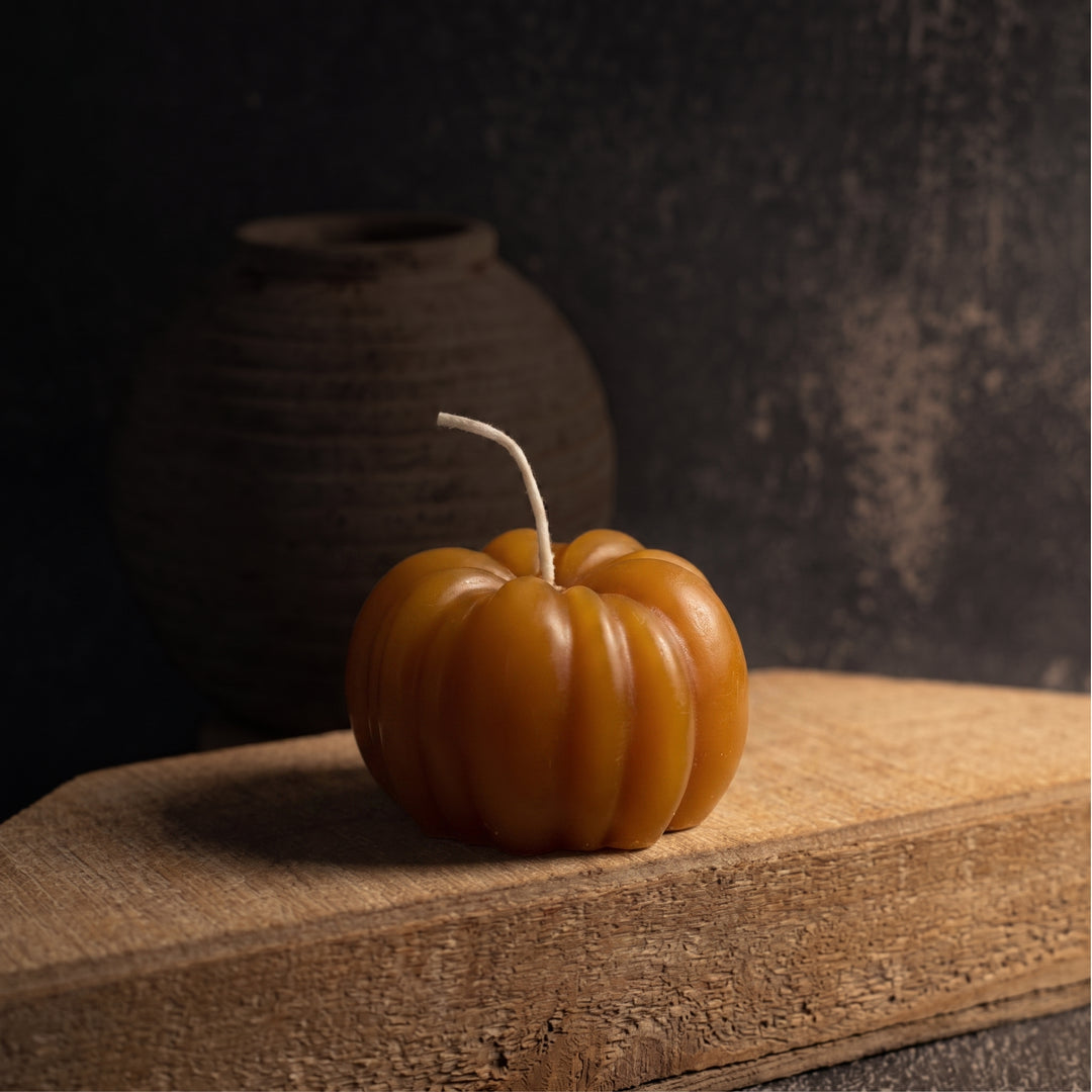 An orange pumpkin-shaped candle with a textured surface and a short white wick, resting on a rustic wooden block against a dark, moody background.