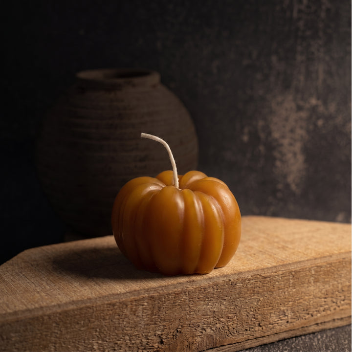 An orange pumpkin-shaped candle with a textured surface and a short white wick, resting on a rustic wooden block against a dark, moody background.