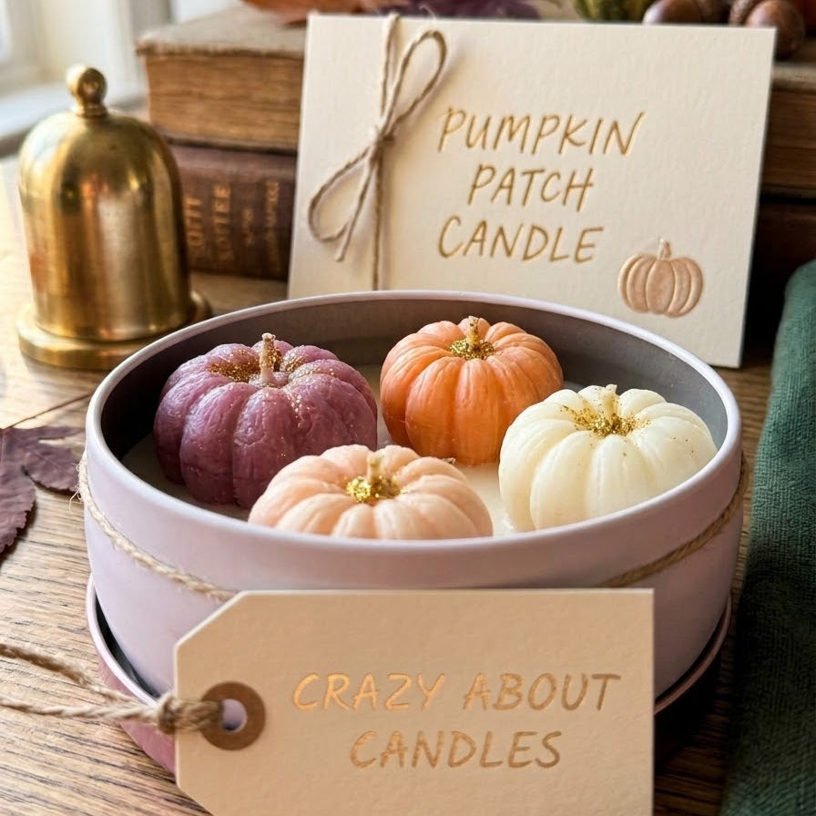 A round pink gift box filled with small decorative pumpkins in purple, peach, pink, and cream, with a card reading 'Pumpkin Patch Candle' and a hanging tag that says 'Crazy About Candles.' The scene is styled on a wooden table with autumn props including books, a brass bell, and more pumpkins in the background.