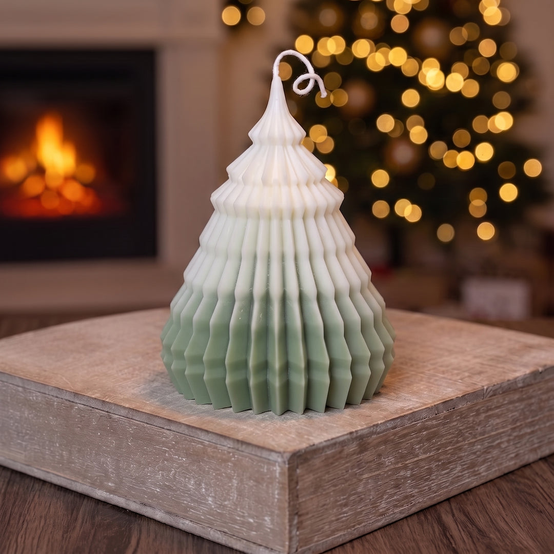 A Christmas tree-shaped candle with a white top fading into a muted sage green base, displayed on a rustic wooden block with a blurred fireplace and festive lights in the background.