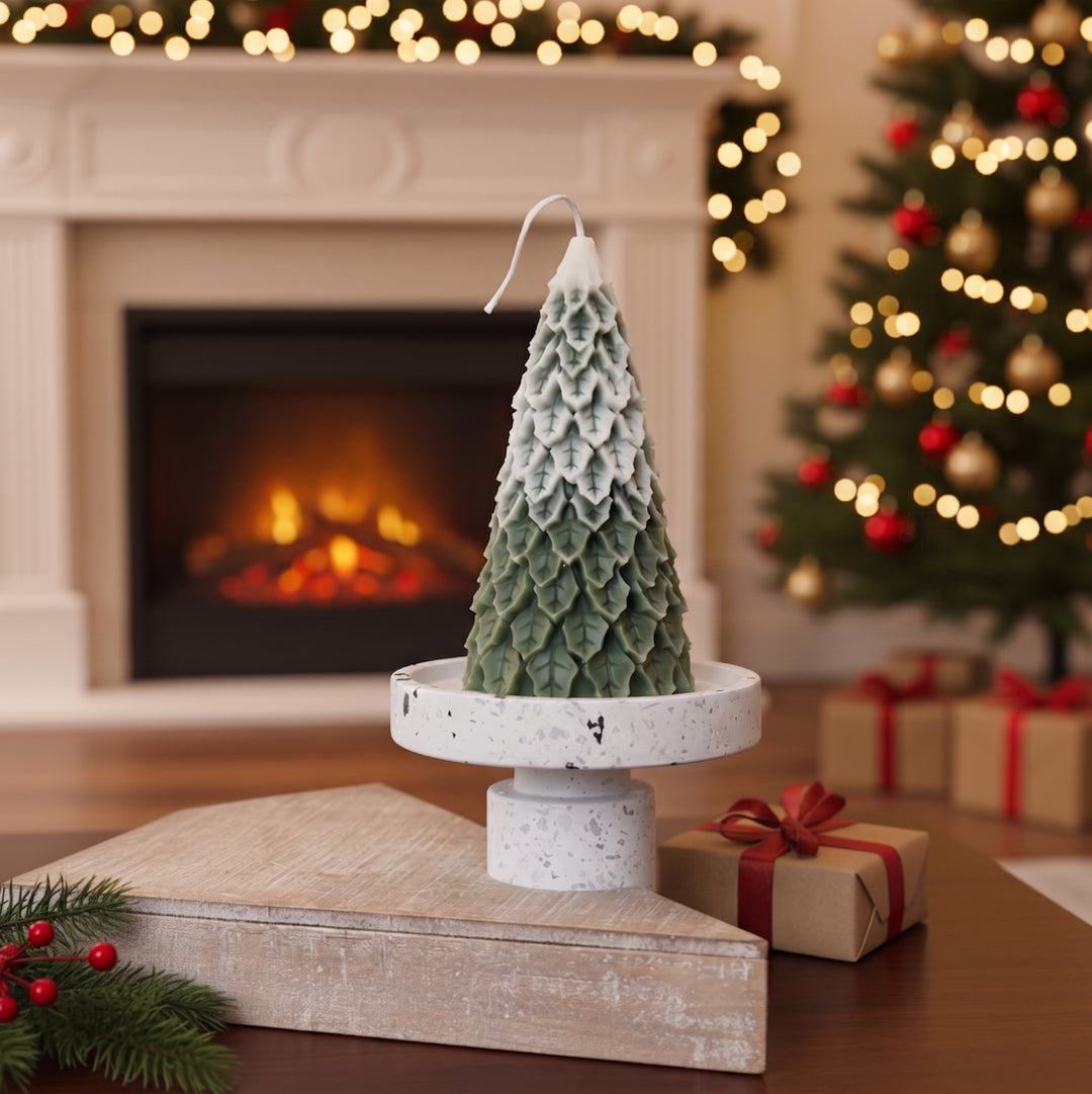 A Large Christmas tree snow capped pine candle with a green and white scalloped pattern sits on a white pedestal stand on a wooden table, with a cozy fireplace, a decorated Christmas tree, and wrapped gifts in the background.