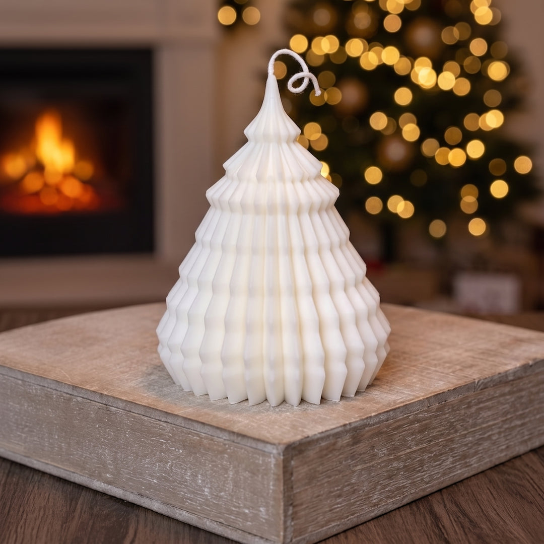 A white Christmas tree-shaped candle with a textured, accordion-style design sits on a rustic wooden block. The background features a warm, blurred fireplace and a glowing Christmas tree with golden bokeh lights.