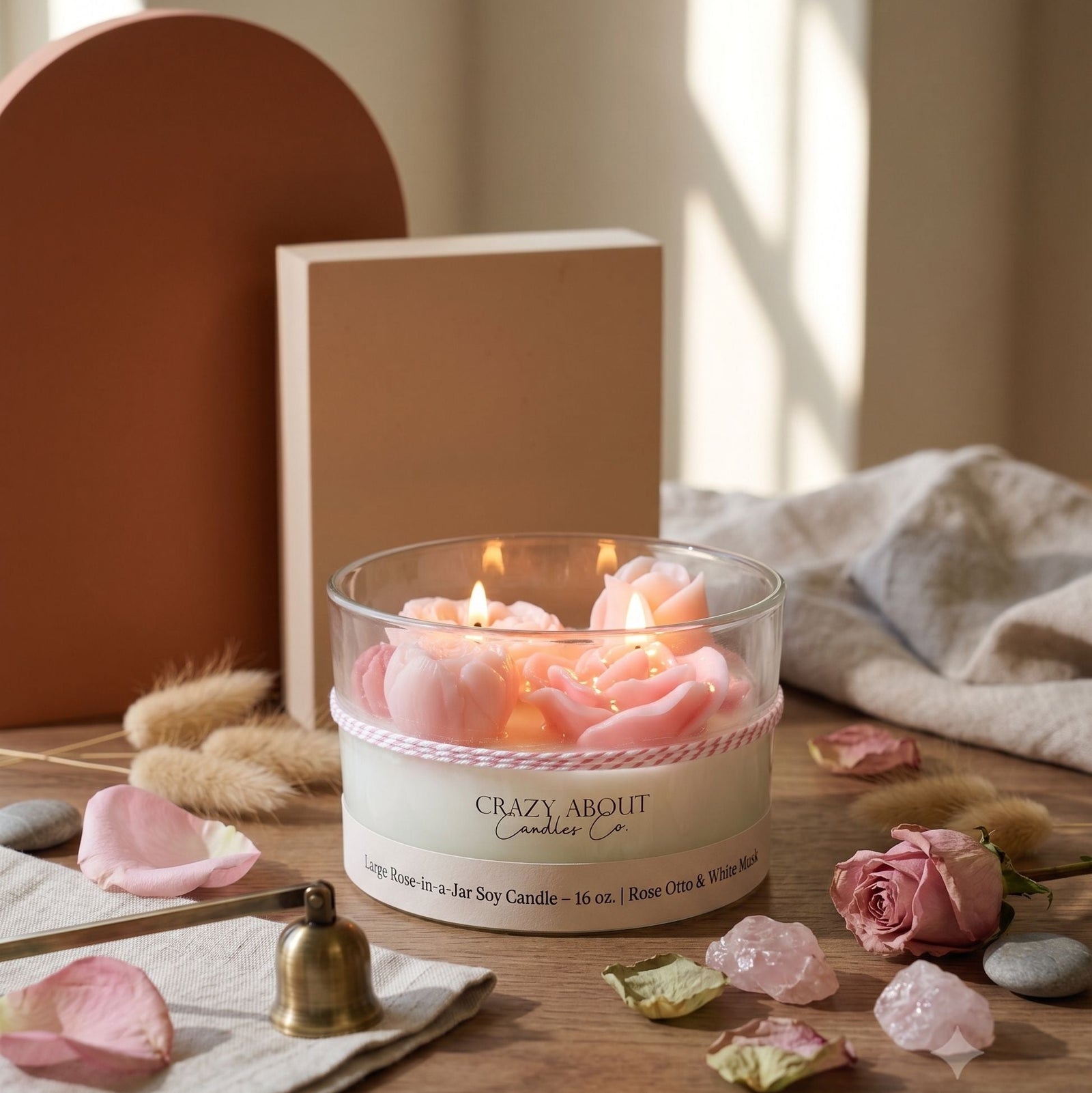A lit jar candle with pink flower-shaped wax pieces inside a clear glass container, placed on a wooden surface with scattered pink petals, crystals, a small bell, and soft home decor in the background. The label reads 'Crazy About Candles Co.
