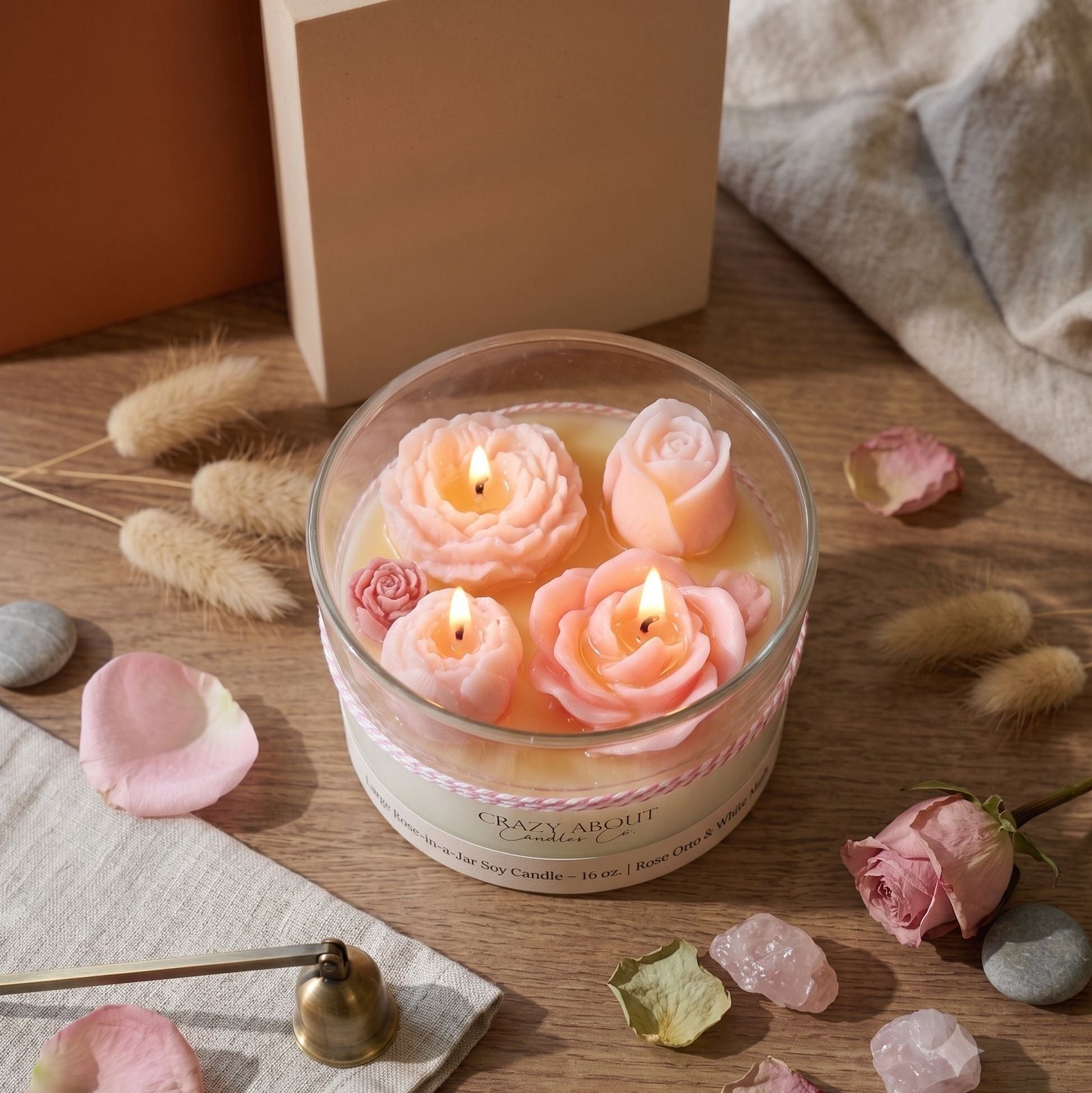 A round glass jar candle with three pink rose-shaped wax candles burning inside, set on a wooden surface with scattered pink petals, dried grass, crystals, and a neutral fabric background. The candle label reads 'Crazy Ambre'.