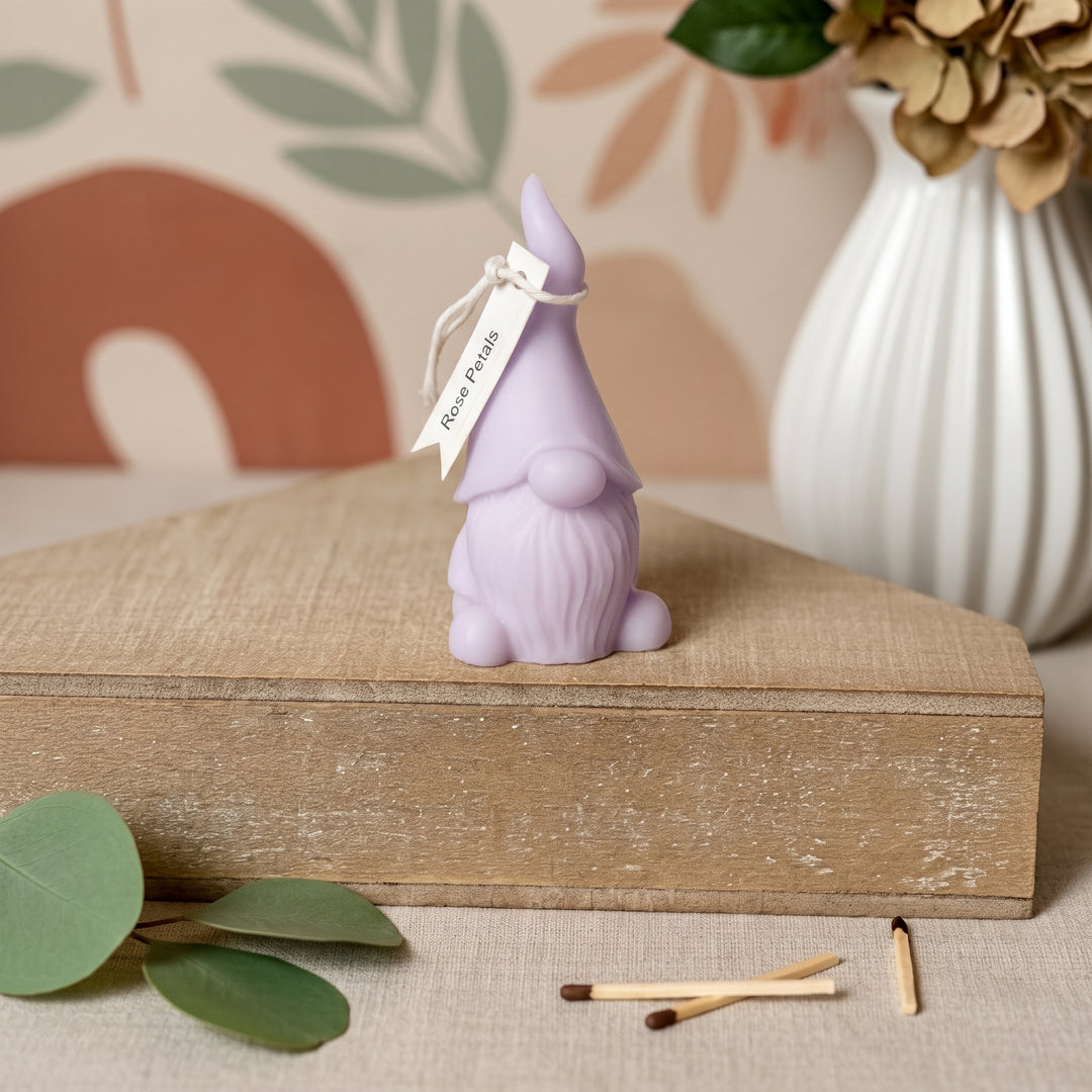 A small lavender gnome figurine with a pointed hat and beard sits on a rustic wooden box. A white paper tag tied to its hat reads &