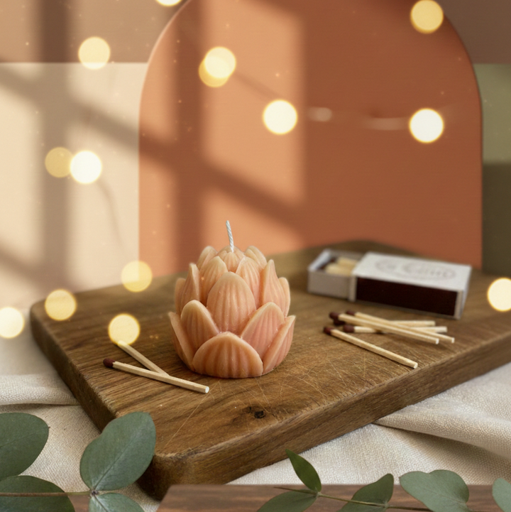 Pink lotus-shaped candle on a wooden board with matches and green leaves in a softly lit room.