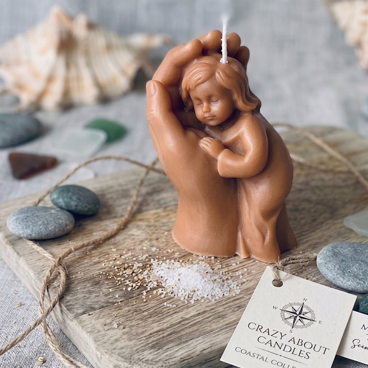 A sculpted candle shaped like a woman cradling a sleeping child, displayed on a rustic wooden board with stones, twine, and a paper tag reading &