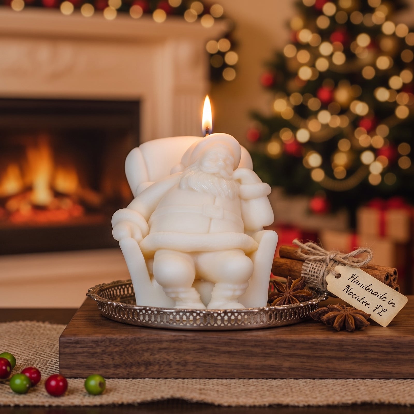 A white Santa Claus seated in an armchair candle burns on a silver tray, styled on a wooden board with cinnamon sticks and a tag that reads 'Handmade in Nevada, Ca.' The background shows a cozy fireplace and a lit Christmas tree in warm, blurred bokeh.